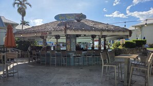 An outdoor seating area with a thatched roof, tables, and chairs. - Sunset Cove 2BR/2BA • Ocean Views • Pool/ Swim-Up Bar • Hot Tub • Walk to Beach! (George Town)