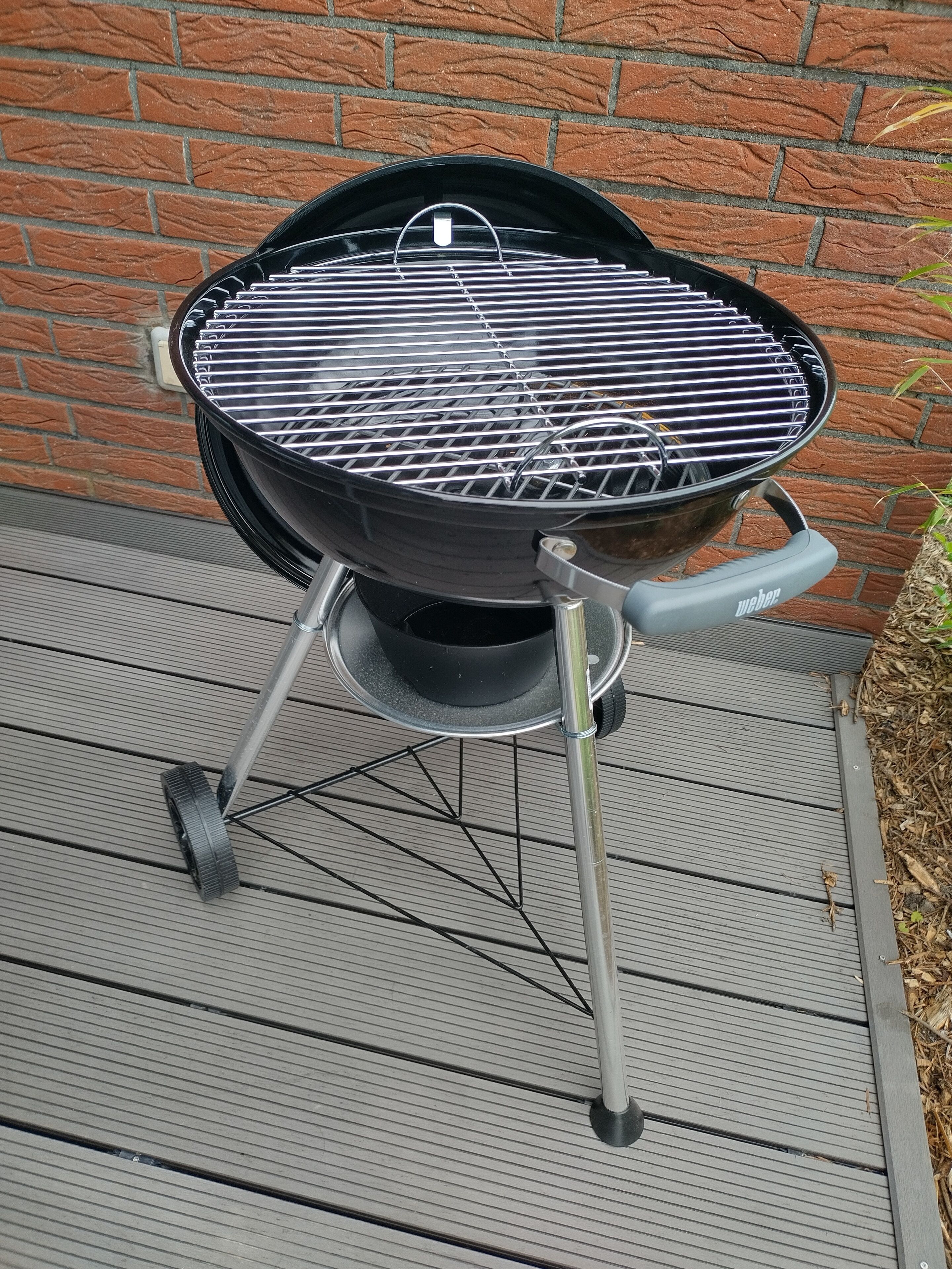 A Weber barbecue grill with a black lid and a handle, placed on a wooden deck with a brick wall in the background.