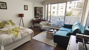 A modern bedroom with a large bed, a desk, a chair, and a sofa. There is a balcony with a view of buildings. - Paris - Porte D'auteuil : Grand Studio Avec Balcon (Paris)