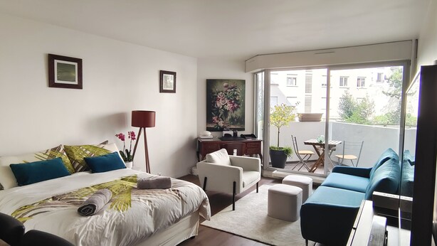 A modern hotel room with a large bed, a desk, a chair, a sofa, and a balcony with a view of the city. - Paris - Porte D'auteuil : Grand Studio Avec Balcon (Paris)