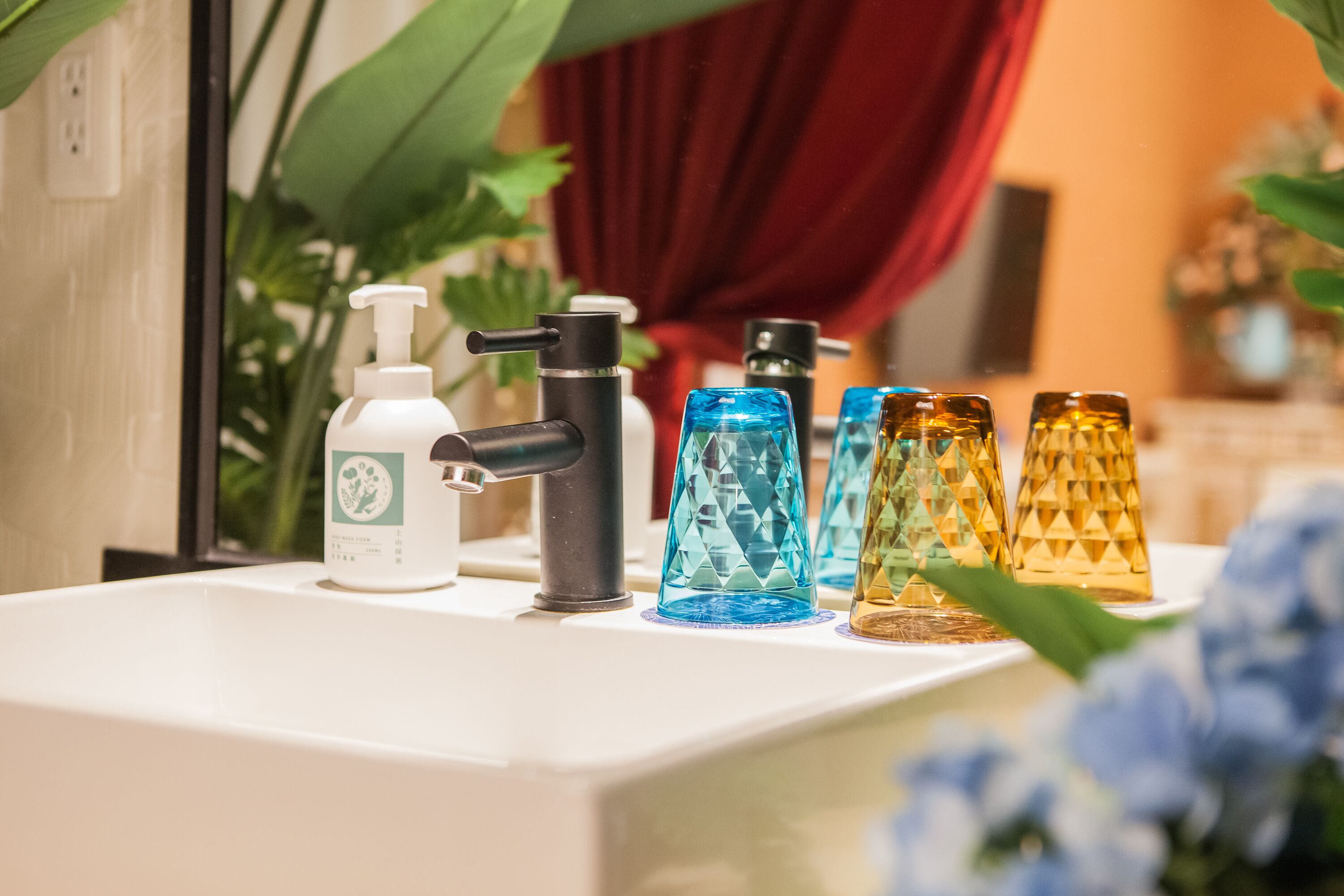 Una encimera de baño con dispensador de jabón, un grifo negro y una fila de recipientes de vidrio de colores.