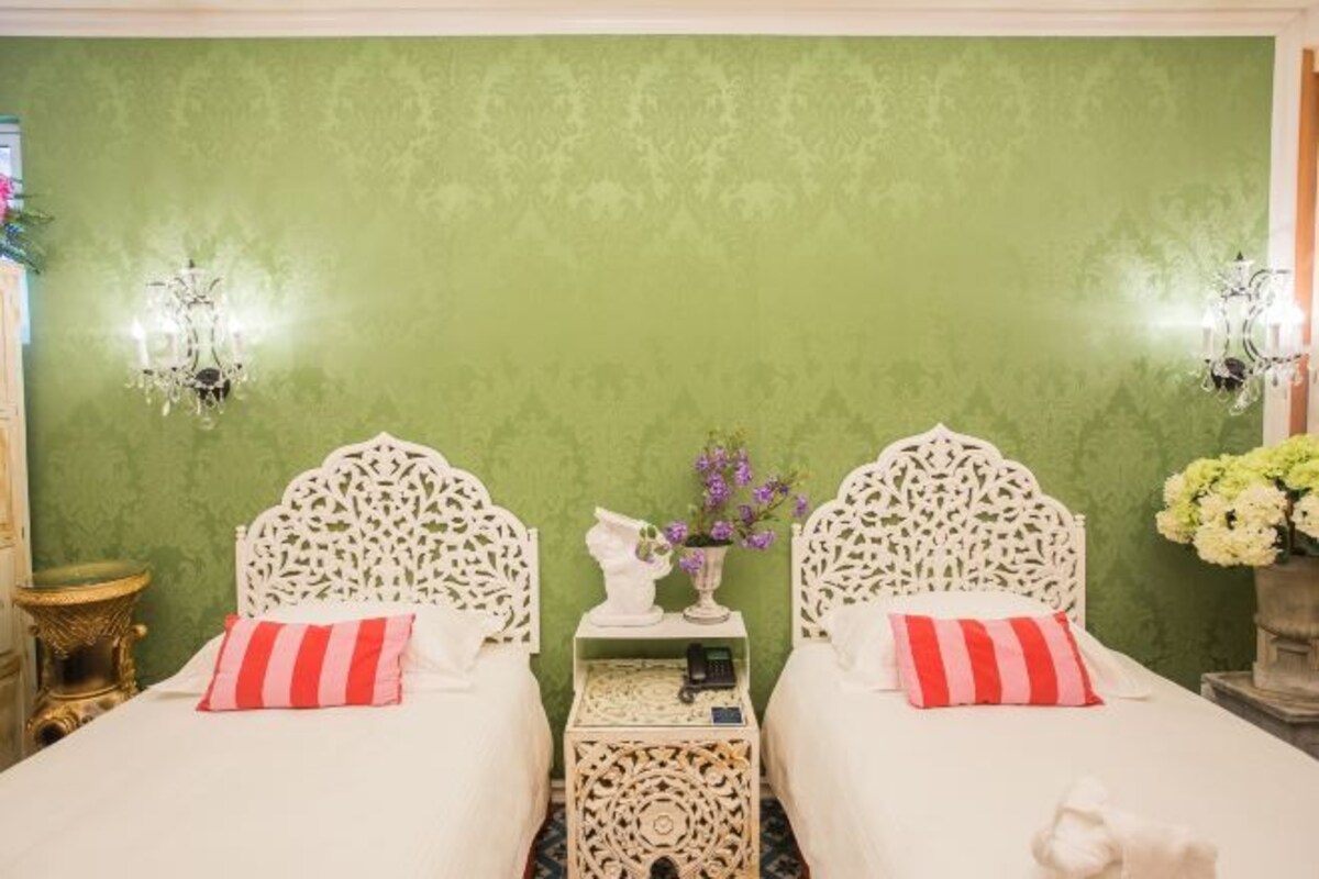 Une chambre avec deux lits, chacun doté d’une tête de lit blanche et d’un oreiller à rayures rouges et blanches. Une petite table se trouve entre les lits, sur laquelle est posé un vase rempli de fleurs violettes.