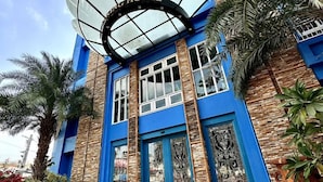 L’entrée d’un hôtel, avec une façade bleue, une grande structure en forme d’éventail au-dessus de la porte et des statues d’éléphants dorées devant.