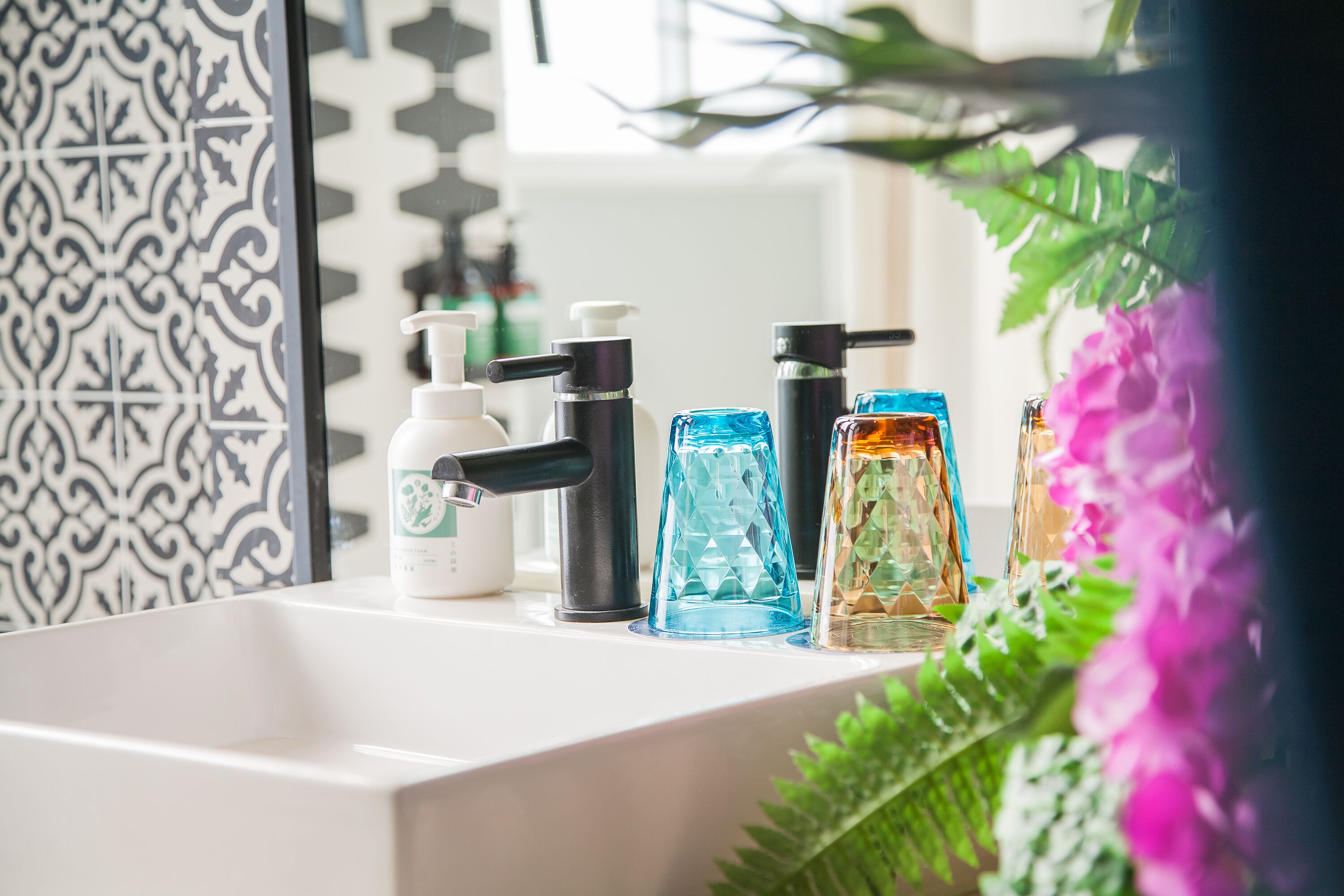 Um banheiro com pia branca, azulejos estampados em preto e branco e diversos produtos de higiene pessoal, incluindo um dispensador de sabonete, um recipiente de vidro azul e um recipiente de vidro transparente.