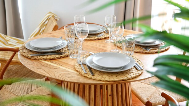 A wooden dining table set with plates, glasses, and cutlery, surrounded by woven placemats and chairs.