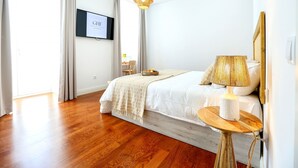 A bedroom with a wooden floor, a bed with a white bedspread, a bedside table with a lamp, and a TV mounted on the wall.
