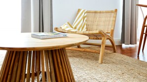 A wicker chair with a striped pillow, a round wooden table with a book, and a woven rug on a wooden floor.