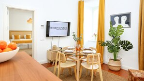 A dining area with a glass table, wooden chairs, a bowl of fruit, a television, and a large plant.