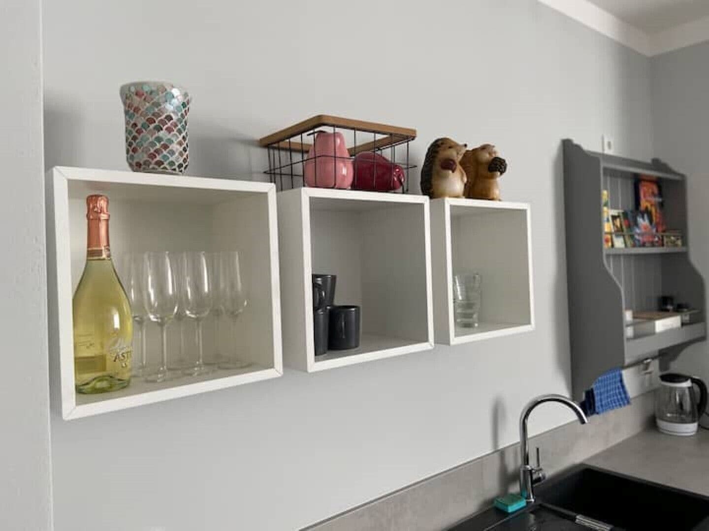 A kitchen with white cabinets, a wine bottle, and glasses on the left shelf, a coffee maker and a kettle on the middle shelf, and a small shelf with decorative items on the right.