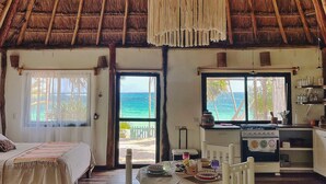 A spacious room with a thatched roof, a dining area with white chairs, a kitchen with a stove, and large windows offering a view of the ocean. - Beautiful cottage in front of the sea. Tulum. (Tulum)