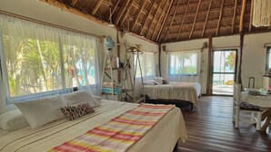 A room with a thatched roof, two beds with white linens, a colorful striped blanket, and a view of palm trees through large windows. - Beautiful cottage in front of the sea. Tulum. (Tulum)