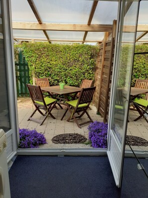En terrasse med et træbord og stole, en grøn potteplante og en hæk i baggrunden.