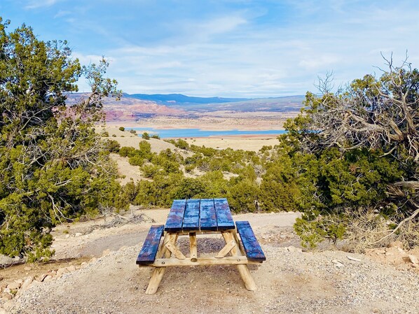 Une table de pique-nique en bois, avec des bancs, située dans un cadre naturel, entourée d’arbres et offrant une vue sur un lac lointain.