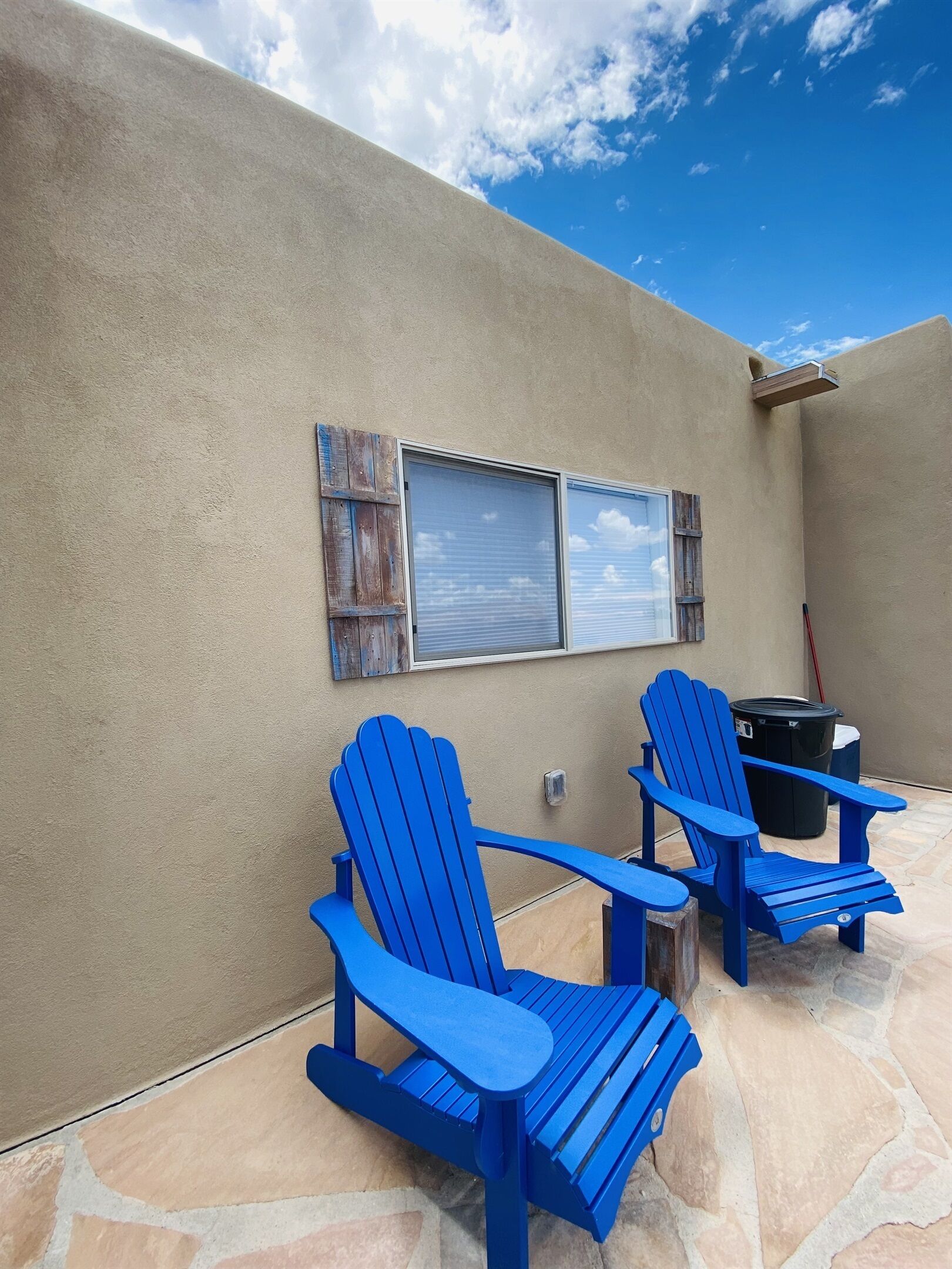 Twee blauwe Adirondack-stoelen op een stenen terras, met een beige muur en een raam met zonwering.