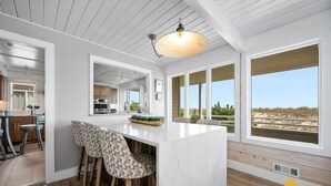 A modern kitchen with a large island, wooden bar stools, and large windows offering a view of the beach.