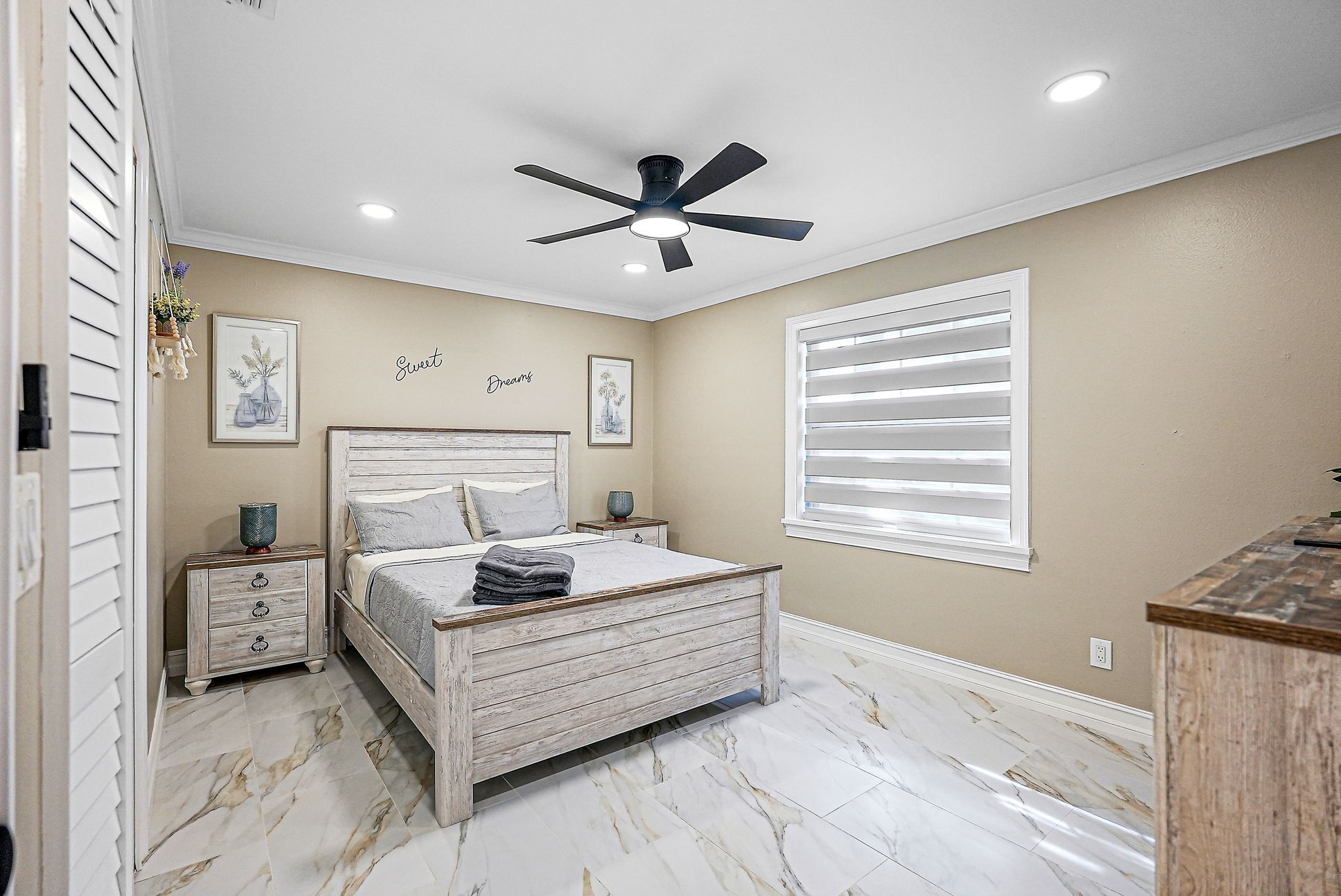 Une chambre à coucher comprenant un lit, un ventilateur de plafond, une table de chevet, une fenêtre avec des stores et un mur orné de tableaux.