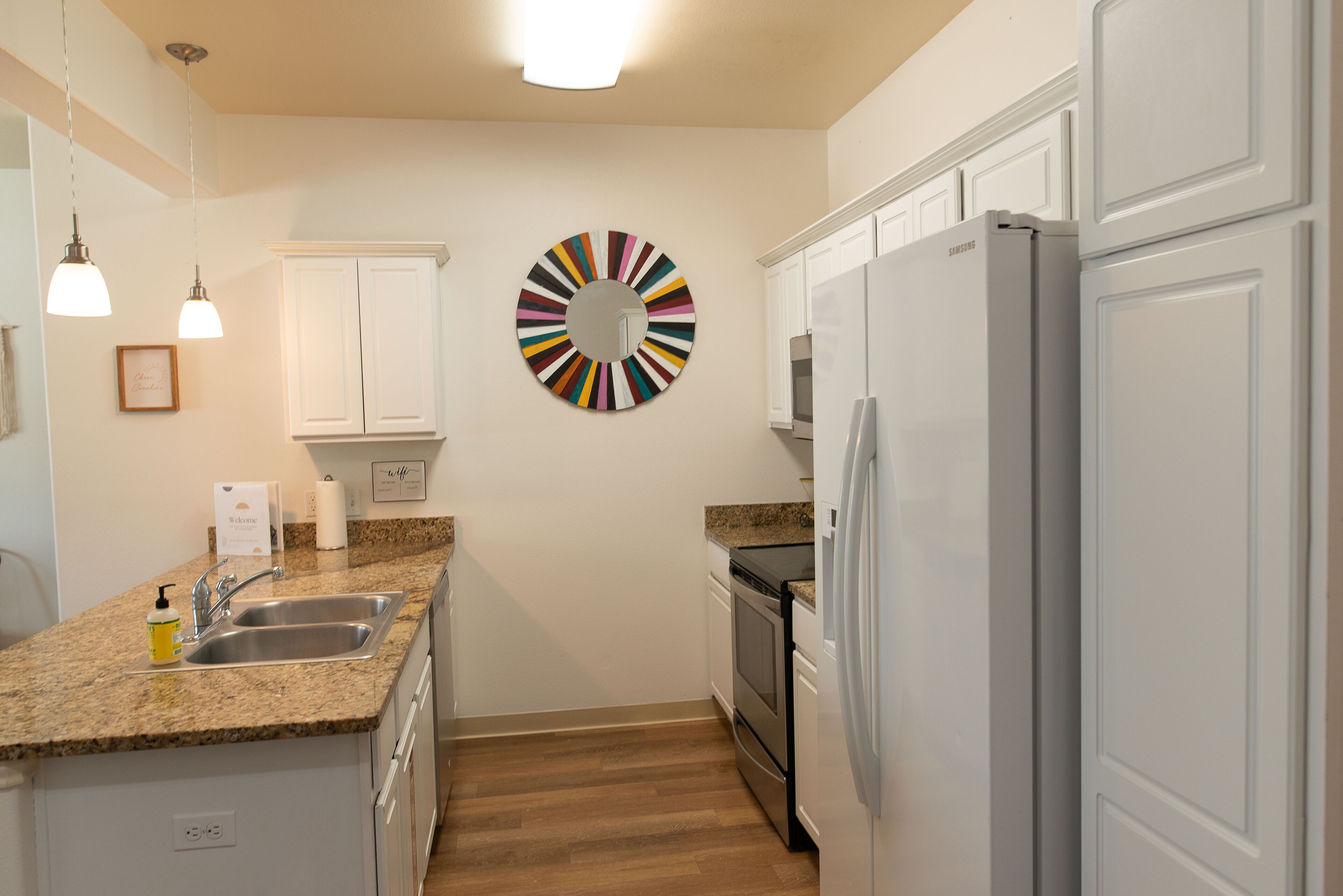 Una cocina con armarios blancos, encimera de granito, electrodomésticos de acero inoxidable y un colorido adorno circular en la pared.
