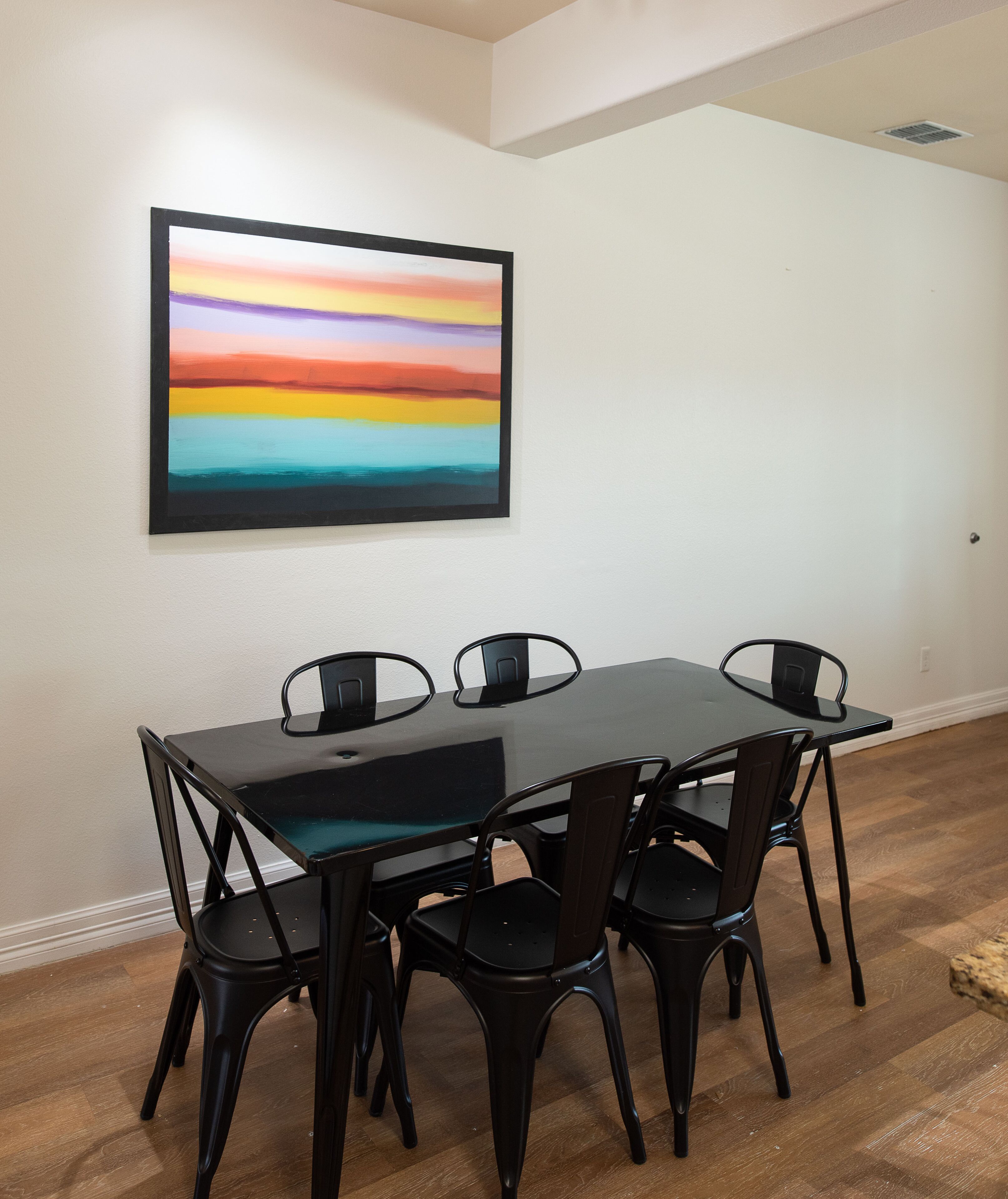 A dining area with a black table and chairs, a colorful painting on the wall, and a wooden floor.