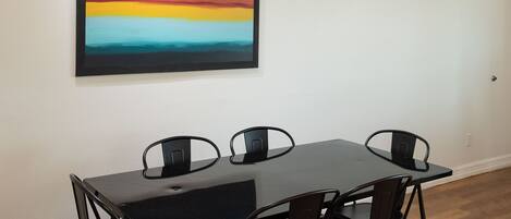 A dining area with a black table and chairs, a colorful painting on the wall, and a wooden floor.