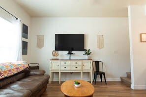 Uma sala de estar com um sofá de couro marrom, uma mesa de centro de madeira, um armário branco, uma cadeira preta e uma TV de tela plana fixada na parede.