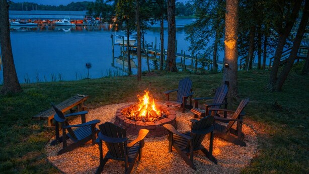 Ein Lagerfeuer mit umlaufenden Stühlen, eine Anlegestelle mit Booten und ein See im Hintergrund.