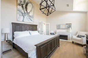 A bedroom with a large bed, a dresser, a chair, a fireplace, and a wall-mounted mirror.