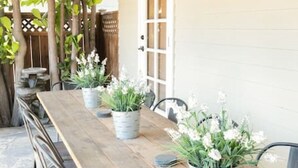 Eine überdachte Terrasse mit einem langen Holztisch, Topfpflanzen und Stühlen.