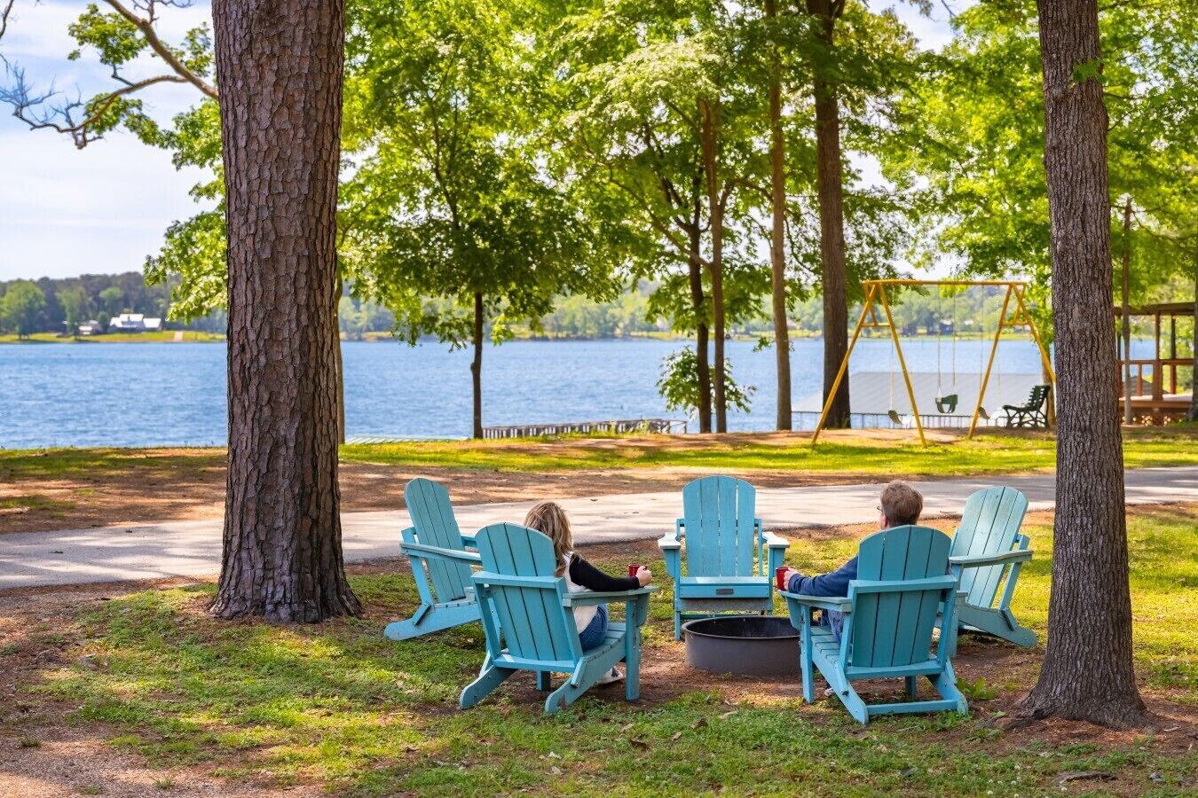 Two people sitting on Adirondack chairs facing a lake, with a swing set and picnic tables in the background.
