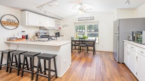 Una cocina moderna con armarios blancos, una isla central con taburetes, una mesa de comedor y vistas al exterior a través de una ventana.