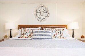 Una cama bien hecha con almohadas estampadas con motivos florales y a rayas, un adorno de pared y dos lámparas de noche.