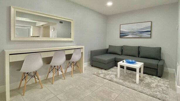 Ein modernes Wohnzimmer mit einem grauen Sofa, einem weißen Schreibtisch mit drei Stühlen, einem großen Spiegel und einem Bild an der Wand.