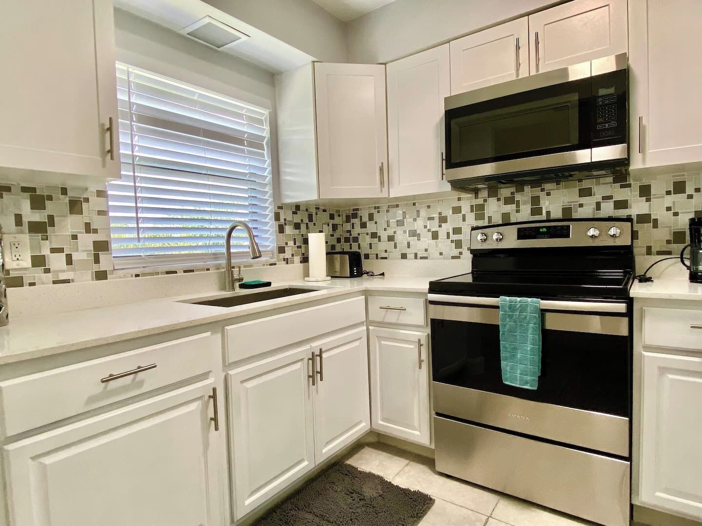 A kitchen with white cabinets, a stainless steel stove, a microwave, and a window with blinds.