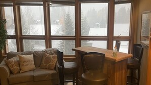 A cozy living room with a sofa, chairs, a wooden table, and large windows overlooking a snowy landscape.