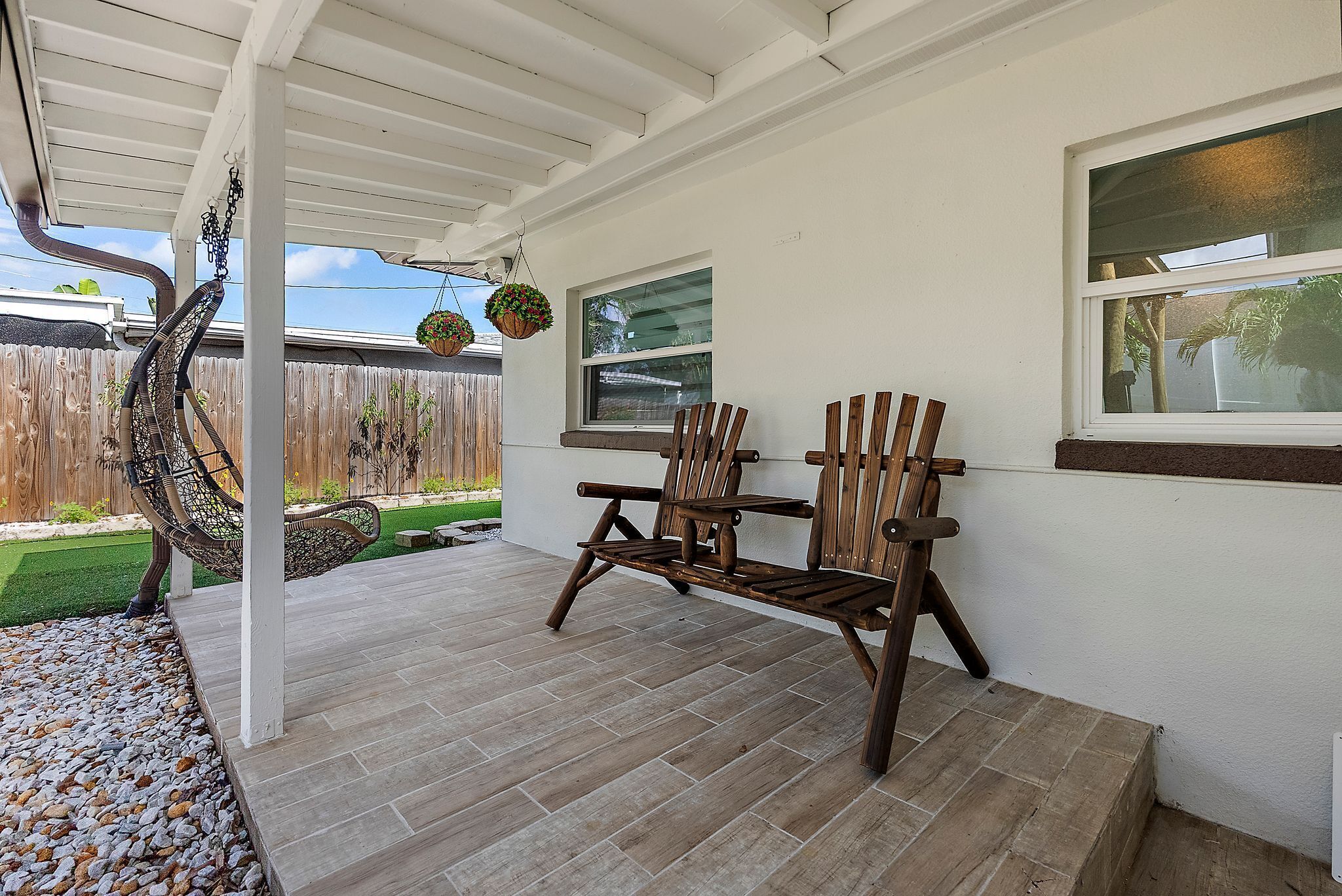 A covered patio with a wooden bench, a hanging chair, and a view of a backyard with a wooden fence and greenery.