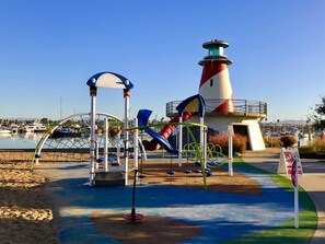 A playground with a slide and climbing structures, a lighthouse, and a marina in the background.