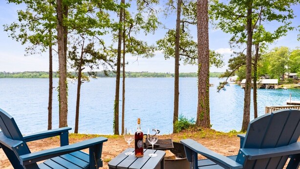 Vista de un lago con dos sillas Adirondack, una mesita y una botella de vino.