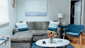 Un salon avec un canapé gris, un fauteuil bleu, une table basse ronde et un tableau représentant des voiliers.