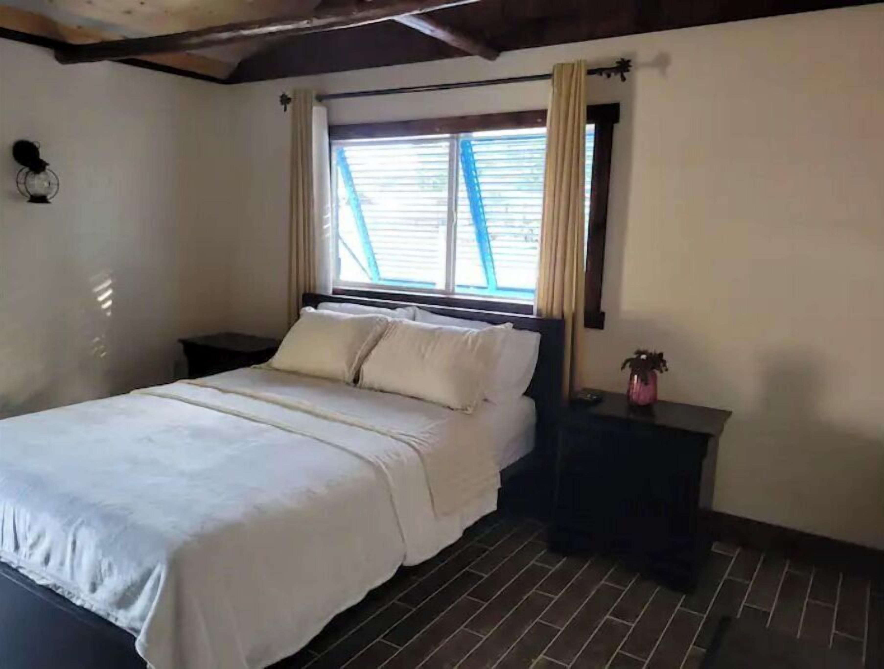A bedroom with a bed, a nightstand, a window with curtains, and a potted plant.