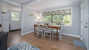 Un comedor con una mesa de madera y sillas blancas, una ventana con vista a árboles y una puerta blanca.