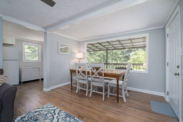 Un comedor con una mesa de madera y sillas blancas, una ventana con vista a árboles y una puerta blanca.