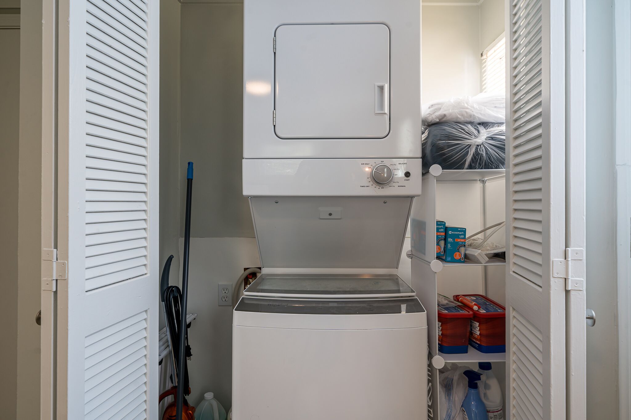 A compact laundry room with a built-in washer and dryer, a mop, and storage shelves with cleaning supplies.