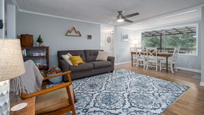 Una sala de estar con un sofá gris, una mesa de comedor de madera, sillas, un tapete estampado y un ventilador de techo.