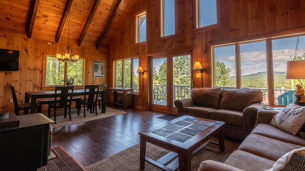 A wooden interior with a dining area, sofa, and large windows offering a view.