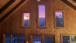 A wooden cabin with a sloped roof, multiple windows, and a view of the sunset through the central window.
