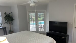Un dormitorio con cama, televisor, ventilador de techo y puerta corrediza de vidrio que da a una zona de piscina.