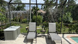 Un porche cubierto con dos sillones de descanso y una pequeña mesa, con vista a una piscina y un jardín.