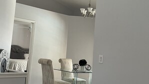 Una sala moderna con una mesa de comedor de vidrio, dos sillones tapizados, una araña y un espejo grande que refleja un dormitorio con cabecera capitonada.