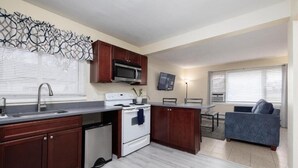 Cozinha compacta com micro-ondas, fogão e lava-louças. Há uma área de refeições com mesa e cadeiras, e um sofá azul na sala de estar.