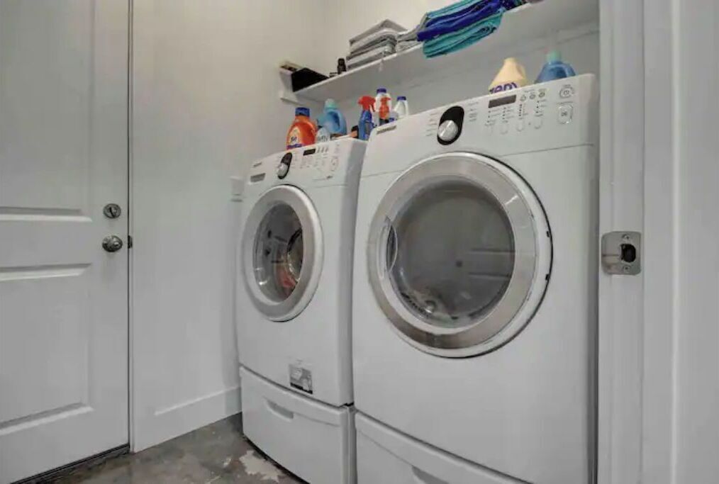 A laundry area with two stacked front-loading washing machines and a shelf above containing various cleaning supplies.