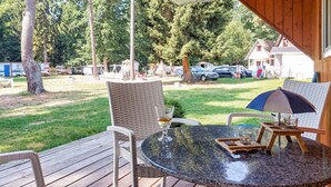 Una terraza de madera con una mesa redonda y sillas, con vistas a un área verde con árboles y coches aparcados.